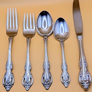 May include: A set of five silver-colored flatware pieces arranged on a yellow surface. The set includes two forks, two spoons, and a knife. Each piece has a decorative floral pattern on the handle.