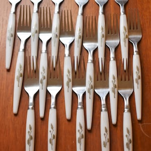 May include: A set of 18 white handled forks with gold floral designs. The forks are arranged in rows on a wooden surface.