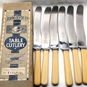 May include: A set of six table knives with light-colored handles and silver blades, displayed next to a vintage cardboard box. The box features text that reads "TABLE CUTLERY" and "JOHN BLYDE LTD." The knives are arranged in a row, showcasing their classic design.