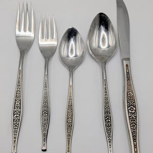 May include: A set of five silver-colored utensils: two forks, two spoons, and a knife. The handles feature a floral design with dark accents. The utensils are arranged on a white surface.