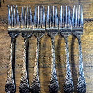 May include: Six silver forks with hammered textured handles. The forks are arranged in a row on a wooden surface.