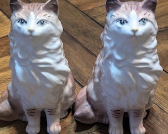May include: Two ceramic cat figurines, one brown and white and the other white and brown, sitting on a wooden surface. Both cats have blue eyes and are looking forward.
