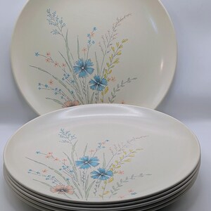May include: A stack of round, off-white dinner plates featuring a delicate botanical illustration of blue cornflowers, yellow wheat stalks, and small pink blossoms. The floral design is centered on the surface of each plate in the set.