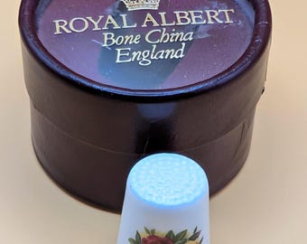 May include: A Royal Albert Bone China thimble from England, displayed next to its original dark maroon box. The thimble is light blue with a floral design in red, yellow, and pink. The box has the brand name and country of origin printed on it.