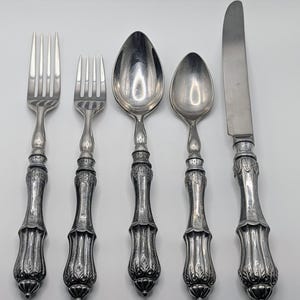 May include: A set of five antique silver-colored utensils, including two forks, two spoons, and a knife. The handles feature an ornate, detailed design. The utensils are arranged on a white surface, showcasing their reflective qualities.