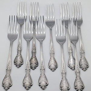 May include: A collection of twelve silver forks. Each fork has a decorative floral pattern on the handle and four tines. The forks are arranged on a white surface.