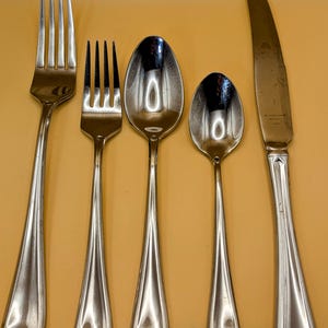 May include: A set of five silver-toned flatware pieces, including a fork, a knife, and three spoons, arranged on a yellow surface.