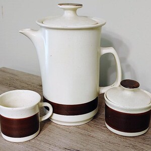 May include: A white ceramic coffee pot with a brown band, a white ceramic creamer with a brown band, and a white ceramic sugar bowl with a brown band.
