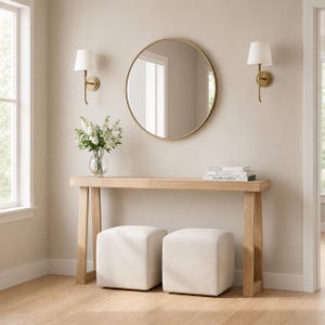 May include: A light wood console table with two matching cream-colored square ottomans underneath. On the table sits a clear glass vase with white flowers and a stack of books. Above the table is a round mirror flanked by two wall sconces.