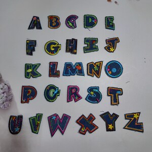 Può includere: Un set completo di ventisei toppe a forma di lettere dell'alfabeto realizzate in tessuto denim blu scuro. Ogni lettera presenta bordi ricamati colorati e cuciture decorative in varie tonalità vivaci, inclusi accenti gialli, rosa, verdi e arancioni.