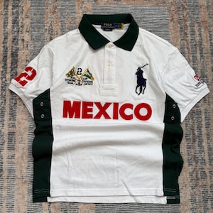 May include: A white short-sleeve polo shirt with a dark green collar and side panels. The front features the word MEXICO in large red letters, a navy blue embroidered polo player logo, and a patch with the text R.R.L. CUP 3 DIVISION PIER MEXICO.