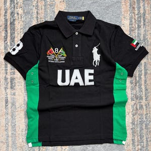 May include: A black short-sleeve polo shirt with green side panels, featuring a white embroidered Polo Ralph Lauren logo, the text UAE, and various nautical-themed patches including the number 8 and a flag of the United Arab Emirates.