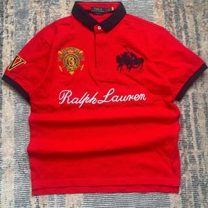 May include: A bright red short-sleeved polo shirt featuring a dark navy collar and sleeve cuffs. The front displays a large white script logo reading Ralph Lauren, a black embroidered polo player graphic, and a circular crest with the number 3.