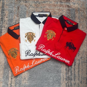 May include: Three short-sleeved polo shirts in orange, white, and red, each featuring a black collar, a circular embroidered crest on the chest, and the text Ralph Lauren in white script embroidery across the lower front section of the fabric.