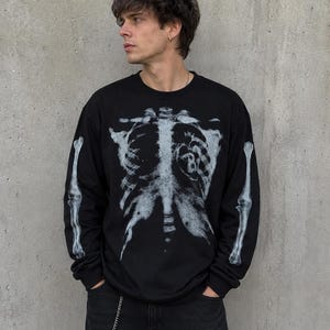 May include: A black long-sleeve crewneck shirt featuring a white, distressed-style graphic of a human rib cage on the front and arm bones printed on each sleeve. The rib cage graphic includes a small heart shape with the number 8 inside it.