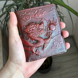 Puede incluir: Una cartera de cuero de color marrón rojizo oscuro con un diseño de dragón en relieve tridimensional. El dragón aparece en una pose dinámica entre nubes estilizadas, con escamas y texturas intrincadas visibles en la superficie del cuero.