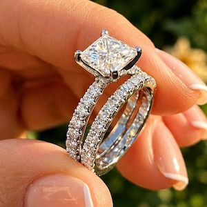 Peut inclure: Une bague de fiançailles argentée avec une pierre centrale taille princesse montée sur quatre griffes, accompagnée d'une alliance assortie. Les deux anneaux sont ornés de petites pierres rondes et transparentes sur toute la surface visible.