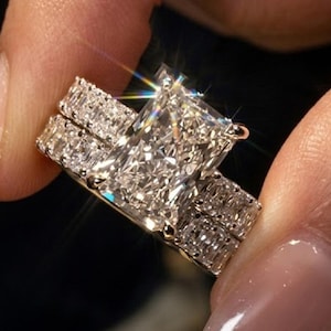 Peut inclure: Une bague de fiançailles en diamant taille radiant avec une grande pierre centrale rectangulaire montée sur un anneau en métal. L'anneau est orné d'une rangée de diamants plus petits taille baguette sur les côtés, créant un aspect étincelant.