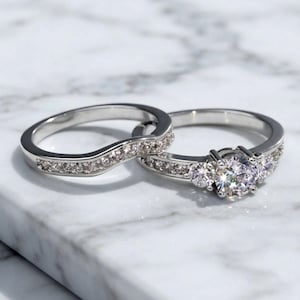 May include: A silver-toned engagement ring set consisting of two pieces. The primary ring features a large central round-cut stone flanked by two smaller stones, while the matching curved band is inlaid with a row of small, sparkling stones.