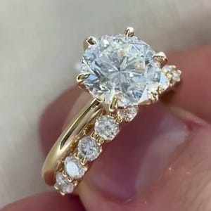 May include: A yellow gold ring featuring a large round brilliant-cut centre diamond held by six prongs. The band is adorned with a row of smaller round diamonds set along the shank, extending partially down the sides of the gold metal band.