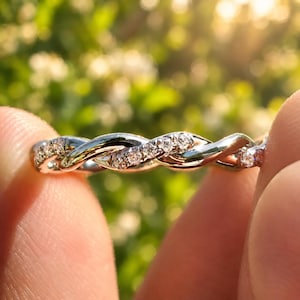 Puede incluir: Un anillo de banda trenzada en tono plateado que presenta una serie de pequeñas piedras transparentes de corte redondo incrustadas a lo largo de uno de los hilos entrelazados. El metal tiene un acabado pulido y reflectante.