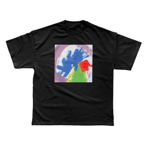 May include: A black short-sleeved t-shirt featuring a square graphic print on the chest. The print displays abstract brushstrokes in vibrant blue, bright green, and red, set against a white background with soft purple accents along the edges.