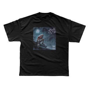 May include: A black short-sleeve t-shirt featuring a square graphic print on the chest. The graphic depicts a person sitting on a ledge in the rain under a streetlamp, with the text Hoodie SZN written in a stylized script above the figure.