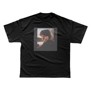May include: A black short-sleeved t-shirt featuring a centered rectangular graphic print. The print displays a photographic image of a person with short brown hair, resting their chin on their hand, set against a backdrop of a snowy landscape.