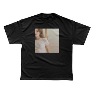 May include: A black short-sleeve t-shirt featuring a centered square graphic print. The print displays a photograph of a person wearing a white t-shirt, with a small white text logo visible in the upper right corner of the square image area.