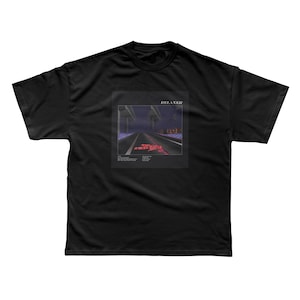 May include: A black short-sleeved t-shirt featuring a central graphic print of a dark road at night under a bridge. The graphic includes the text RELAXER at the top and smaller text blocks below the road, with a red abstract shape on the asphalt.