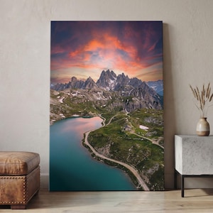 Può includere: Una stampa su tela verticale che raffigura un paesaggio montano con un lago turchese, sentieri sterrati tortuosi e una piccola baita immersa in colline verdi. Le vette frastagliate e innevate si stagliano contro un cielo al tramonto drammatico con vivaci nuvole arancioni e viola.