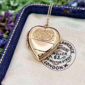 May include: A gold heart-shaped locket pendant suspended on a delicate chain. The locket features an engraved floral pattern on the upper half and a smooth, polished finish on the lower half, resting inside a blue velvet-lined jewelry box.
