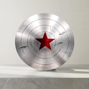 Puede incluir: Un escudo circular met&aacute;lico con acabado en plata cepillada que presenta una estrella roja prominente en el centro. La superficie muestra ranuras circulares conc&eacute;ntricas, detalles de l&iacute;neas grabadas y dos marcas de ara&ntilde;azos horizontales.