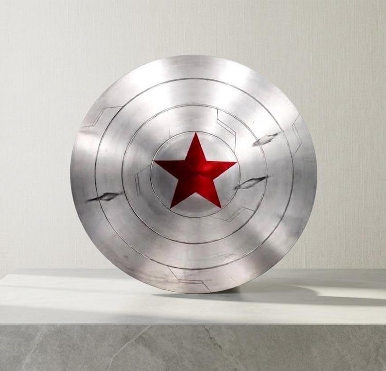 Puede incluir: Un escudo circular met&aacute;lico con acabado en plata cepillada que presenta una estrella roja prominente en el centro. La superficie muestra ranuras circulares conc&eacute;ntricas, detalles de l&iacute;neas grabadas y dos marcas de ara&ntilde;azos horizontales.