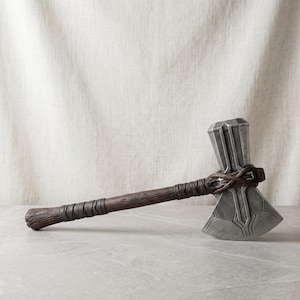 May include: A battle axe with a metallic silver head featuring intricate geometric engravings and a dark brown wooden handle textured to resemble bark, secured at the head with dark brown leather-like wrappings.