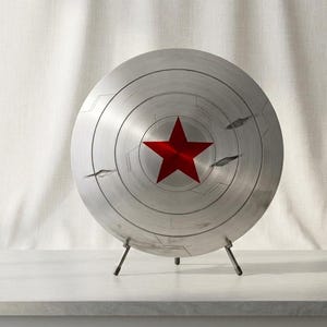 Puede incluir: Un escudo circular plateado con una estrella roja prominente en el centro. La superficie met&aacute;lica muestra ranuras circulares conc&eacute;ntricas y varias marcas de impacto o hendiduras oscuras y dentadas sobre la cara del escudo.