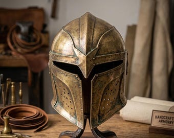 Puede incluir: Un casco de metal en tono latón desgastado con una visera puntiaguda, una ranura horizontal para los ojos y paneles laterales perforados. El casco está montado sobre un soporte de metal oscuro con una base de madera circular y acabado rústico.