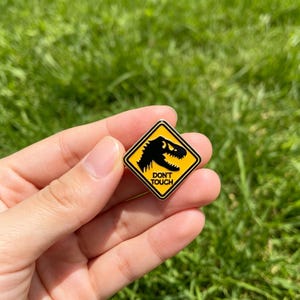 May include: A diamond-shaped enamel pin featuring a yellow background with a black border and a black silhouette of a dinosaur head. The text DON'T TOUCH is printed in black capital letters below the dinosaur graphic on the pin surface.