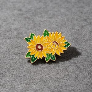 Op de afbeelding: Een metalen emaille speld met twee gele zonnebloemen met bruine harten en groene bladeren. Het ontwerp is omlijnd met goudkleurig metaal en toont een paar bloeiende bloemen met gedetailleerde bloemblaadjes en bladeren als een kleurrijk accessoire.