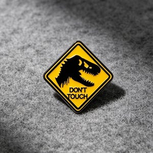 May include: A diamond-shaped enamel pin featuring a yellow background with a black border. The design includes a black silhouette of a dinosaur head and the text DON'T TOUCH in black capital letters, mimicking a yellow road warning sign.