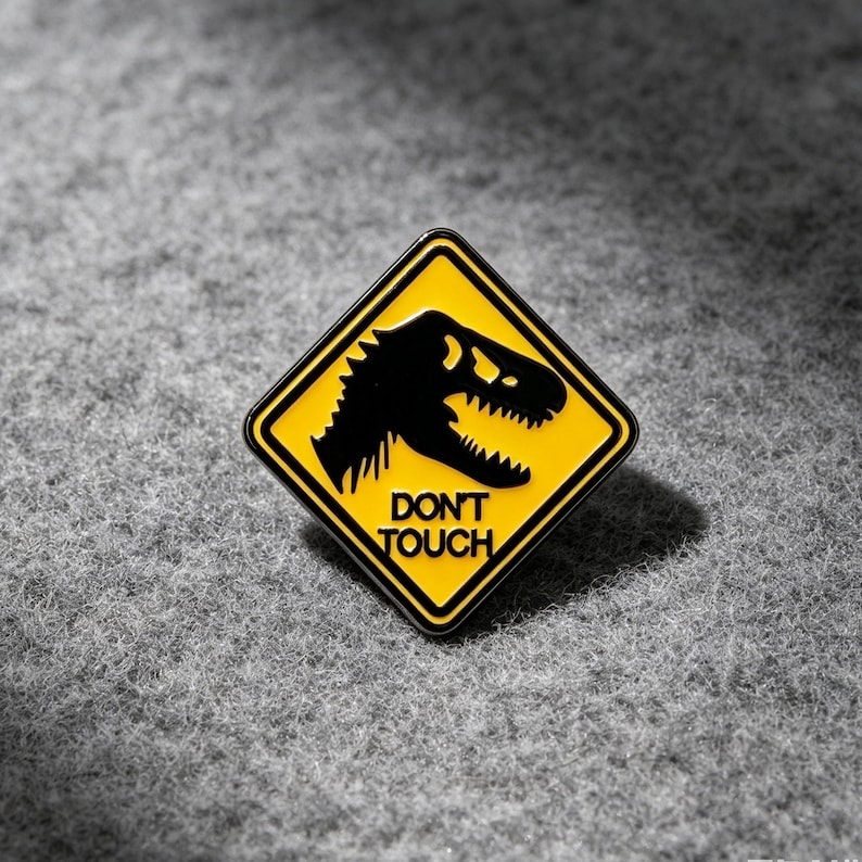 May include: A diamond-shaped enamel pin featuring a yellow background with a black border. The design includes a black silhouette of a dinosaur head and the text DON'T TOUCH in black capital letters, mimicking a yellow road warning sign.