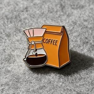 May include: A metal enamel pin featuring a pour-over coffee maker with a light pink filter and dark brown liquid, positioned next to an orange coffee bag with the word COFFEE printed in black lettering, all outlined in a gold-toned metal finish.