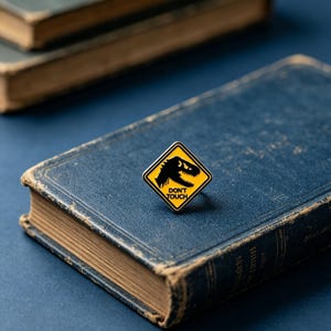 May include: A diamond-shaped enamel pin featuring a yellow background with a black border and a black silhouette of a dinosaur head. The text DON'T TOUCH is printed in black capital letters below the dinosaur graphic on the pin surface.