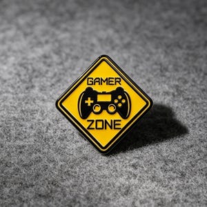 Op de afbeelding: Een ruitvormige emaille speld met een gele achtergrond en een zwarte rand. Het ontwerp bevat een zwarte afbeelding van een videogamecontroller, gecentreerd tussen de woorden GAMER en ZONE in dikke, zwarte, schreefloze hoofdletters.