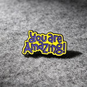 May include: A metal enamel pin featuring a bright yellow background with a black border and the text "You are Amazing!" written in bold, purple, rounded lettering. The design is shaped to follow the contours of the text.