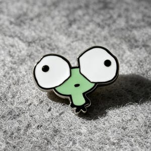 Puede incluir: Un pin de esmalte metálico con forma de cabeza de rana de dibujos animados con dos ojos blancos grandes y sobredimensionados con pupilas negras, una cara de color verde claro y una boca pequeña negra, delineado con un borde de metal negro.