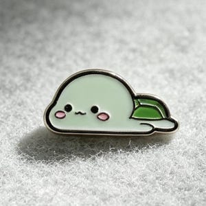 Puede incluir: Un pin de esmalte metálico con forma de pequeña tortuga de color verde claro con un caparazón verde oscuro. La tortuga tiene un cuerpo liso y redondeado, dos ojos de punto negro, una boca diminuta y dos pequeños círculos rosas en las mejillas, todo delineado en metal negro fino.