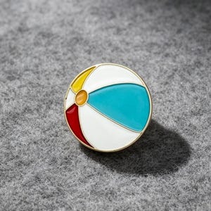 May include: A round enamel pin featuring a beach ball design with a gold-toned metal border. The beach ball graphic is divided into segments coloured in white, bright turquoise, yellow, and red, with a small orange circle at the centre point.
