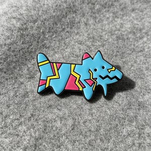 Op de afbeelding: Een metalen emaille speld in de vorm van een gestileerde hond. De hond is lichtblauw met een grillig, geometrisch patroon in felroze en geel. Het heeft een zwarte omlijning, twee kleine zwarte stippen als ogen en een zigzaglijn als mond.