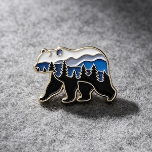 Può includere: Una spilla in smalto con bordo dorato a forma di orso, caratterizzata da un design paesaggistico a strati. L'interno raffigura una sagoma nera di pini su una catena montuosa blu, sormontata da un cielo bianco e una piccola luna circolare, il tutto bordato d'oro.
